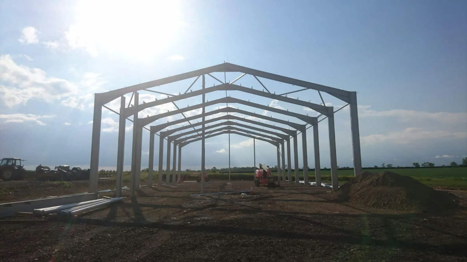 Bare steel portal frame silhouetted against a bright sky, with purlins in place and open fields beyond