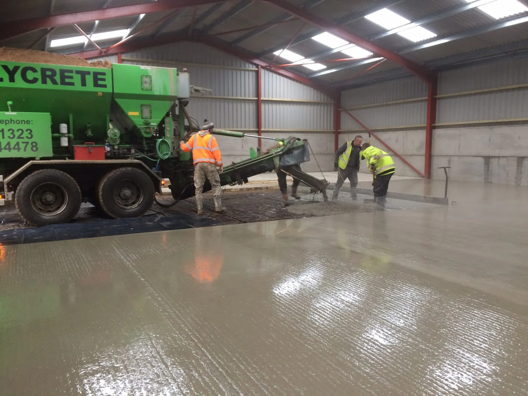 Team in hi-vis power-floating a wet concrete slab inside a red-framed building, with a volumetric mixer truck alongside