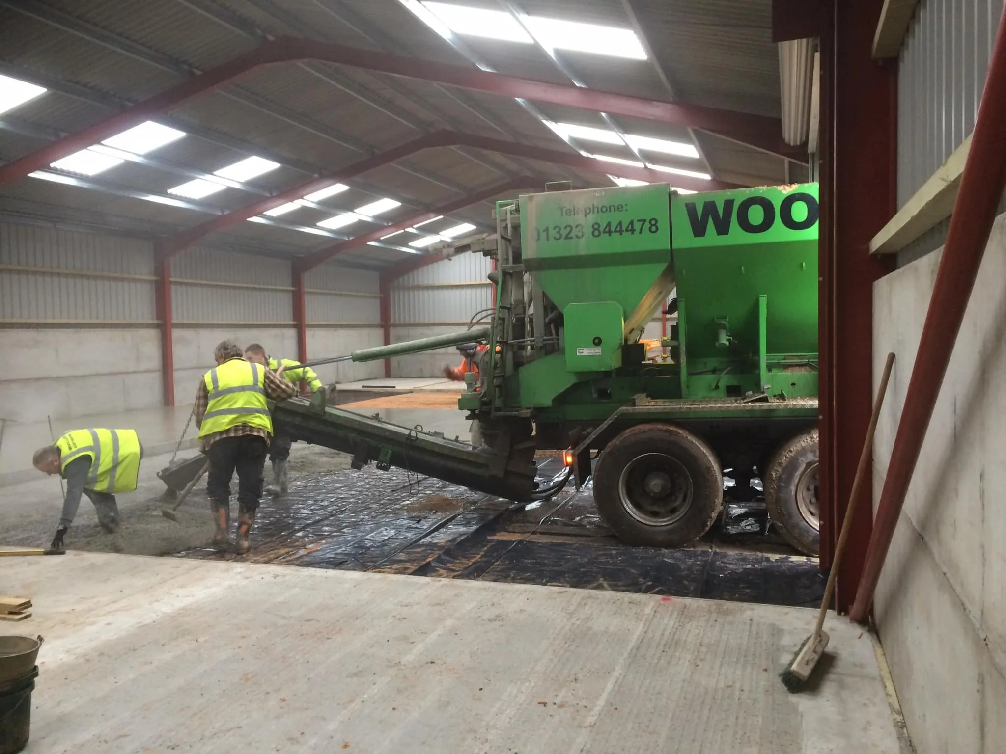 Green volumetric concrete mixer truck discharging into a chute while workers screed the slab