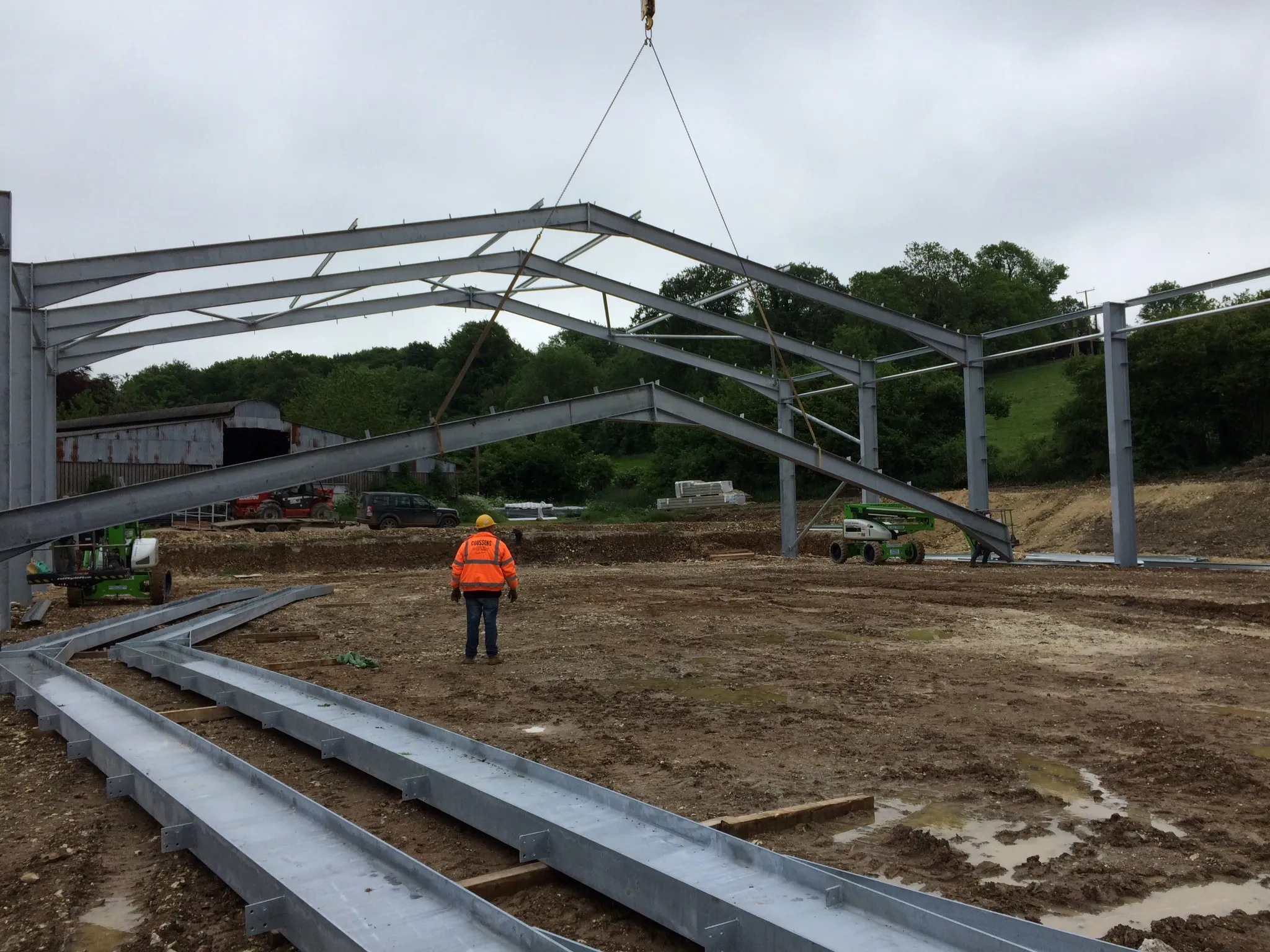 Site worker in hi-vis guiding a long steel beam suspended from a crane above a prepared base