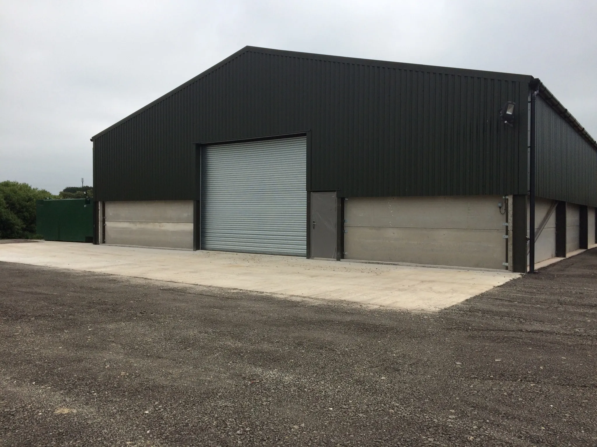 Dark green machinery shed with two roller shutter doors and a wide concrete hardstanding